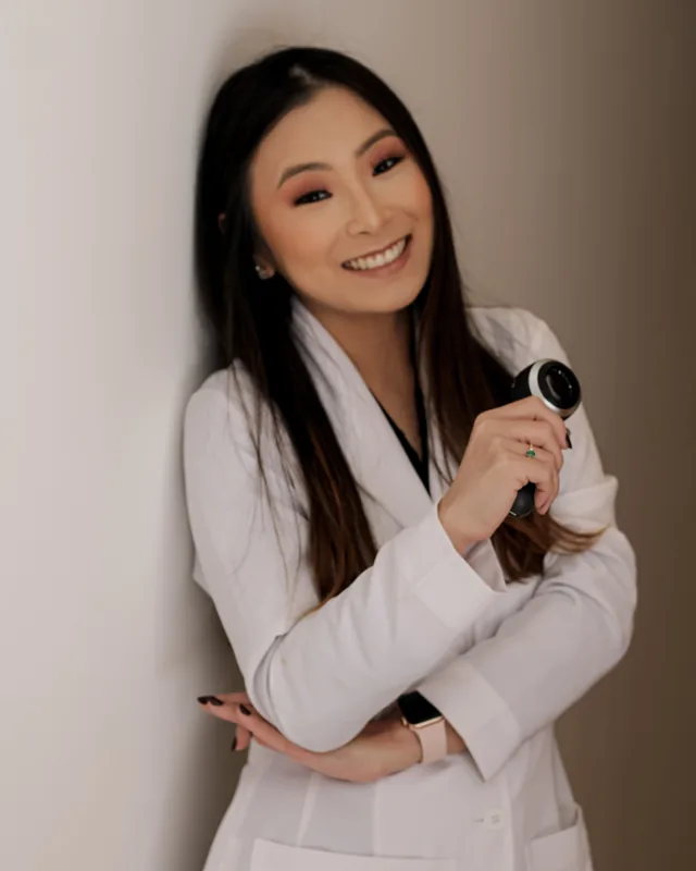 Dra. Maísa Okada de jaleco, sorrindo, com um dermatoscópio na mão.